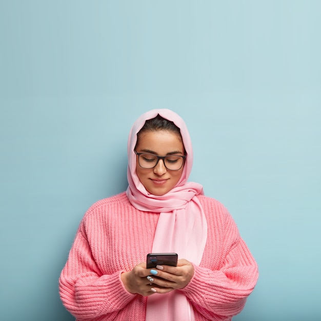 Charmante, vriendelijk ogende vrouw in hijab, volgt de islam, kijkt aandachtig naar het schema van morgen, gebruikt moderne mobiele telefoon, zoekt informatie op internet, draagt een oversized trui, geïsoleerd op blauwe muur