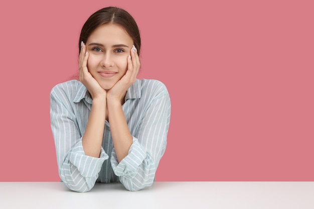 Charmante vriendelijke jonge blanke vrouw in stijlvolle kleding zittend aan een bureau met de handen op haar gezicht, met belangstelling, gelukkig lachend. Menselijke gezichtsuitdrukkingen en lichaamstaal