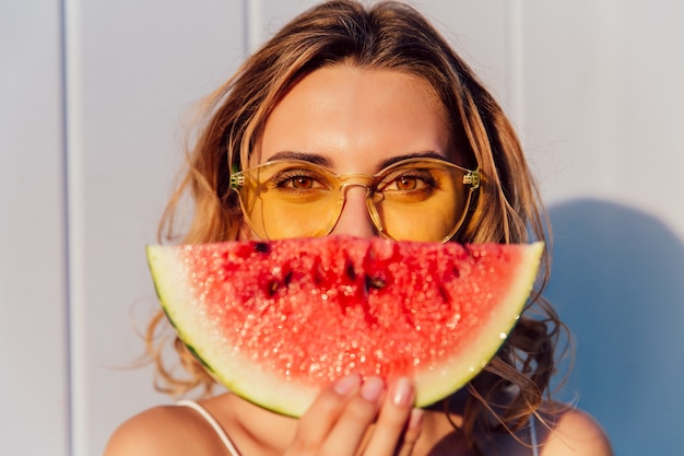 Charmante vrouw die in gele zonnebril de helft van haar gezicht met stuk van watermeloen verbergt