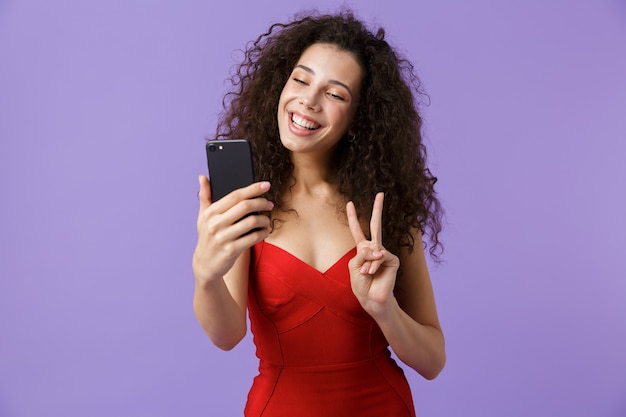 Charmante vrouw, gekleed in rode jurk met zwarte smartphone, geïsoleerd over violette muur
