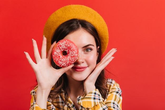 Charmante vrouw in baret poseren met donut op rode muur. meisje in geel overhemd is schattig lachend.