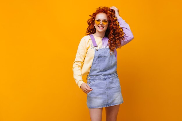 Charmante vrouw in blauwe jumpsuit en bril lacht op oranje muur