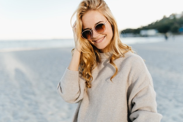 Charmante vrouw met golvend blind haar, gekleed in lichte trui en zonnebril met glimlach tegen zee.