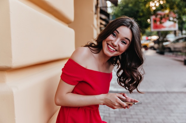 Charmante vrouw met golvend haar staande in de buurt van de telefoon te bouwen en vast te houden. donkerharige blithesome meisje in een rode jurk lachen naar de camera.