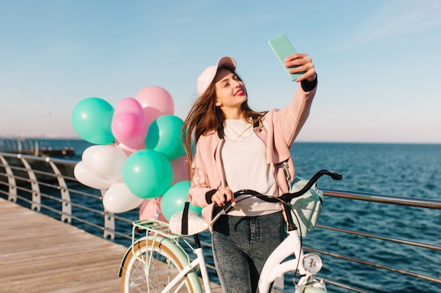 Charmante vrouwelijke wielrenner in een roze pet en windjack glimlachend en selfie nemen op de achtergrond van de zee. Aanbiddelijk donkerbruin meisje met witte fiets en kleurrijke partijballons die pret naast oceaan hebben.