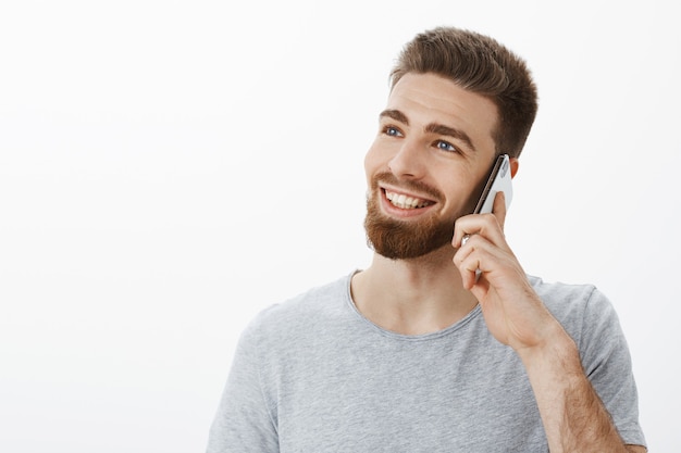 Charmante, zorgeloze jonge succesvolle ondernemer met blauwe ogen en baard die mobiel in de buurt van oor houdt en naar links kijkt met een dromerige, vrolijke blik, glimlachend terloops pratend via smarpthone over witte muur