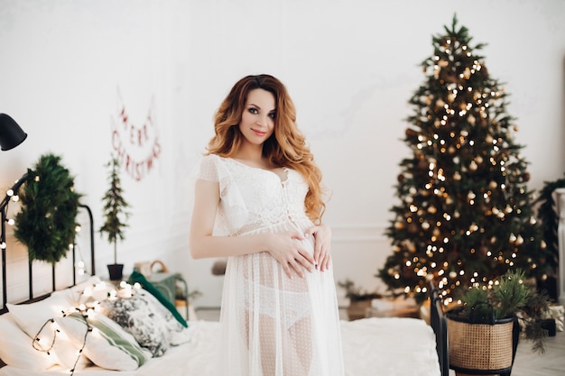 Charmante zwangere vrouw poseert voor de camera in witte jurk in de buurt van kerstboom met een lor van lichten