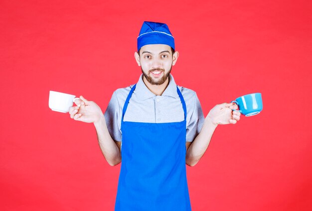 Chef-kok in blauwe schort met blauwe en witte keramische kopjes in beide handen.