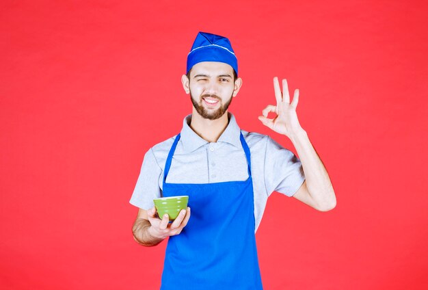 Chef-kok in blauwe schort met een groene keramische beker en tevredenheidsteken.