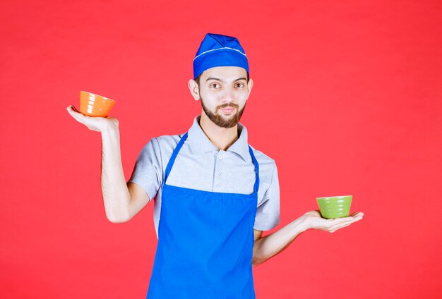 Chef-kok in blauwe schort met gele en groene keramische kopjes in beide handen.