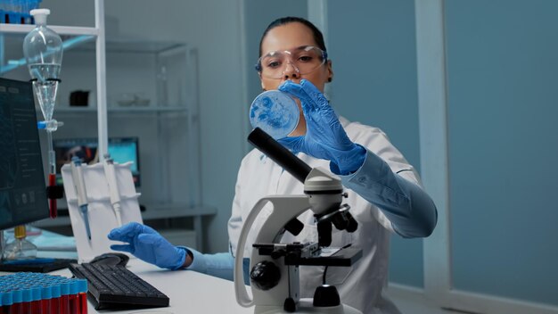 Chemiewetenschapper die onderzoeksapparatuur in laboratorium op bureau met petrischaal, micropipet, vacutainers en microscoop gebruikt. Wetenschappelijke vrouw die op computer typt voor ontwikkeling