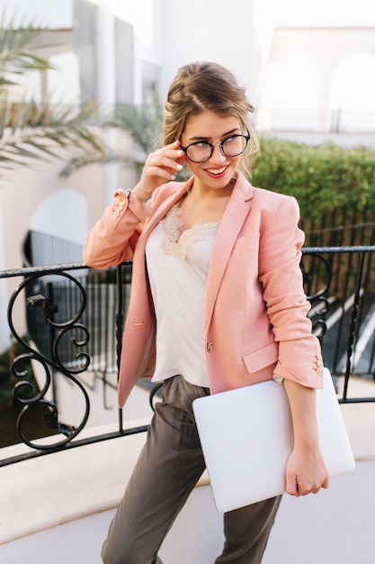 Cherful jonge zakenvrouw, student met laptop in de hand, staande op mooi balkon, terras in hotel, restaurant, resort. Modieuze bril, roze jasje, beige blouse, grijze broek.