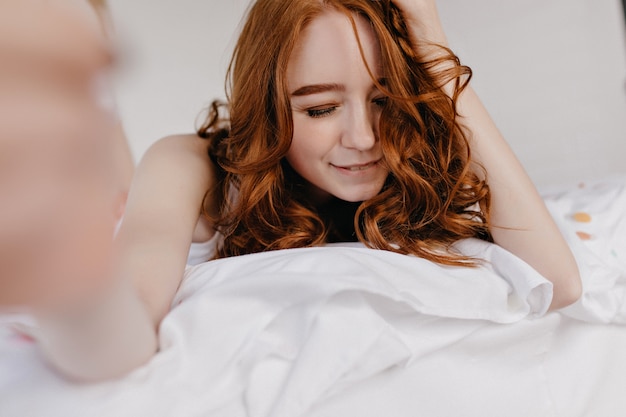 Chillen gember vrouw selfie maken in de ochtend. Verfijnd Kaukasisch vrouwelijk model dat een foto van zichzelf neemt terwijl ze in haar kamer ligt.