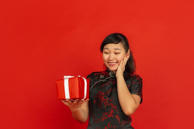 Chinees Nieuwjaar 2020. Het portret van het Aziatische jonge meisje dat op rode achtergrond wordt geïsoleerd. Vrouwelijk model in traditionele kleding ziet er blij, lachend en verrast uit door een geschenkdoos. Viering, vakantie, emoties.