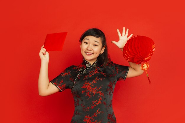 Chinees Nieuwjaar 2020. Het portret van het Aziatische jonge meisje dat op rode achtergrond wordt geïsoleerd. Vrouwelijk model in traditionele kleding ziet er blij uit met decoratie en rode envelop. Viering, vakantie, emoties.