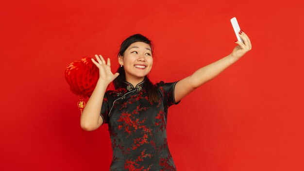 Chinees Nieuwjaar 2020. Het portret van het Aziatische jonge meisje dat op rode achtergrond wordt geïsoleerd. Vrouwelijk model in traditionele kleding ziet er gelukkig uit en neemt selfie met decoratie. Viering, vakantie, emoties. Folder.