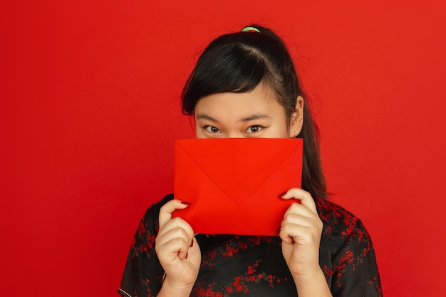 Chinees nieuwjaar. het portret van het aziatische jonge meisje dat op rode achtergrond wordt geïsoleerd. vrouwelijk model in traditionele kleding ziet er dromerig uit en toont rode envelop. viering, vakantie, emoties.