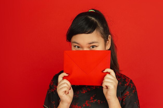 Chinees Nieuwjaar. Het portret van het Aziatische jonge meisje dat op rode achtergrond wordt geïsoleerd. Vrouwelijk model in traditionele kleding ziet er dromerig uit en toont rode envelop. Viering, vakantie, emoties.