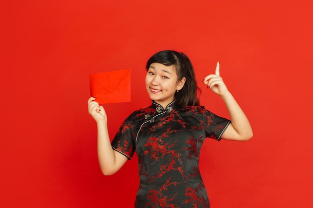 Chinees Nieuwjaar. Het portret van het Aziatische jonge meisje dat op rode achtergrond wordt geïsoleerd. Vrouwelijk model in traditionele kleding ziet er gelukkig uit, glimlachend en wijzend op rode envelop. Viering, vakantie, emoties.