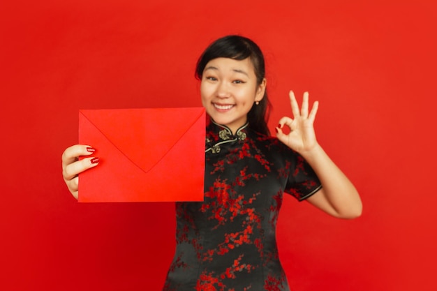 Gratis foto chinees nieuwjaar. het portret van het aziatische jonge meisje dat op rode achtergrond wordt geïsoleerd. vrouwelijk model in traditionele kleding ziet er gelukkig uit, lacht en toont rode envelop. viering, vakantie, emoties.