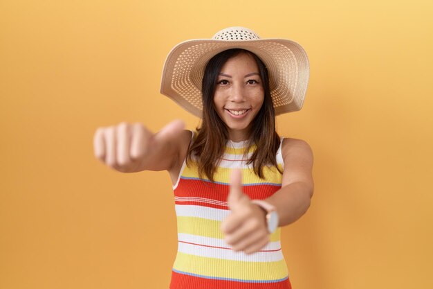 Chinese vrouw van middelbare leeftijd met zomerhoed op gele achtergrond die positief gebaar met de hand goedkeurt, duimen omhoog glimlachend en blij voor succes. winnaar gebaar.