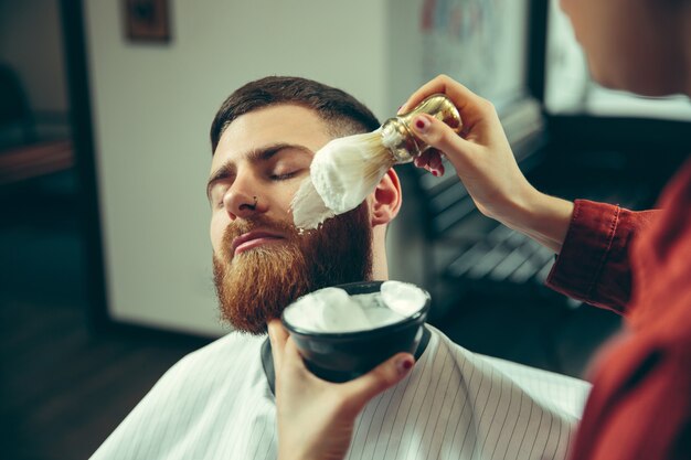 Cliënt tijdens baardscheren in kapperszaak. Vrouwelijke kapper bij salon. Geslachtsgelijkheid. Vrouw in het mannelijke beroep.