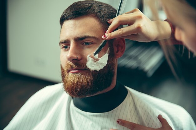 Cliënt tijdens baardscheren in kapperszaak. Vrouwelijke kapper bij salon. Geslachtsgelijkheid. Vrouw in het mannelijke beroep.