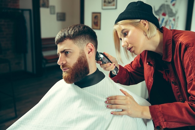 Cliënt tijdens het scheren van de baard in de kapperszaak. Vrouwelijke kapper bij salon. Geslachtsgelijkheid. Vrouw in het mannelijke beroep.