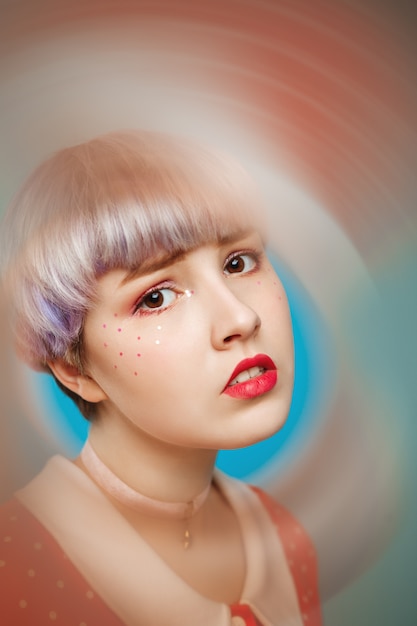 Close-up artistiek conceptueel portret van mooi popachtig meisje met kort licht violet haar, gekleed in rode jurk over blauwe muur Onscherpe voorgrond.