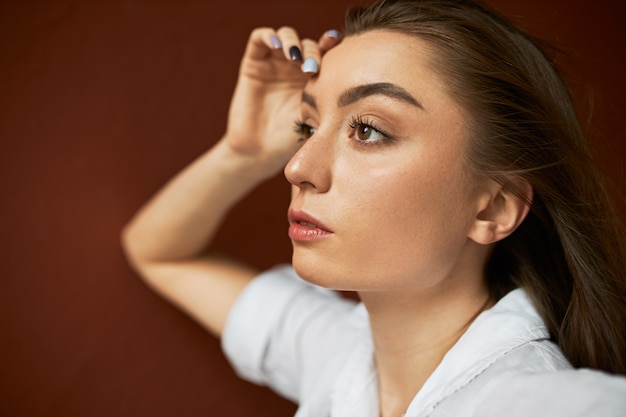 Close-up beeld van aantrekkelijke doordachte jonge blanke vrouw met schone huid en los bruin haar gezicht aanraken, peinzende gelaatsuitdrukking hebben, vooruitkijkend, iets herinnerend