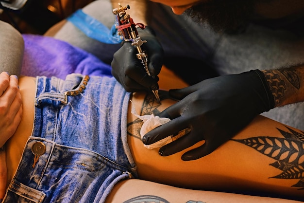 Close-up beeld van de bebaarde mannelijke tatoeëerder maakt een tatoeage op een vrouwelijk been.