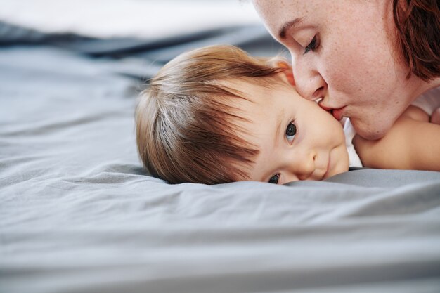 Close-up beeld van de jonge moeder die haar dochtertje op de wang kust als ze na het spelen op bed rust