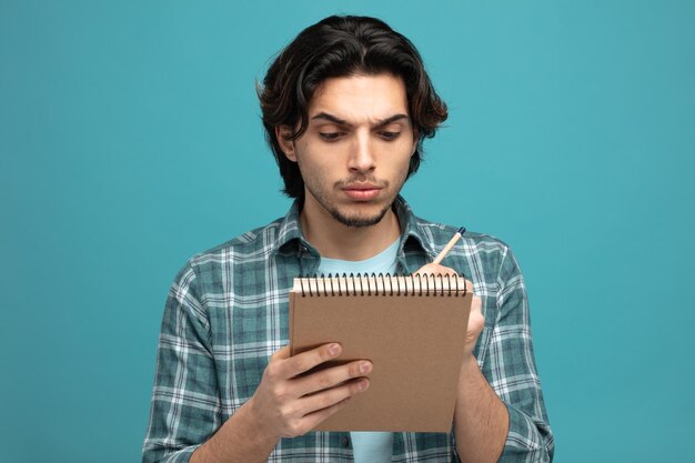 close-up beeld van geconcentreerde jonge knappe man die iets schrijft met potlood op notitieblok geïsoleerd op blauwe achtergrond