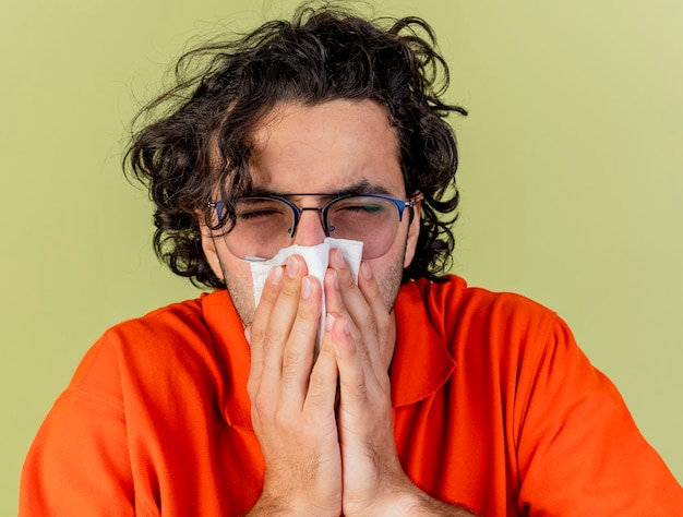 Gratis foto close-up beeld van jonge zieke man met bril neus afvegen met servet met gesloten ogen geïsoleerd op olijfgroene muur