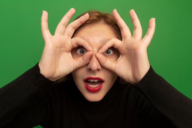 Gratis foto close-up beeld van onder de indruk jonge blonde vrouw die naar voren kijkt en een gebaar doet met handen als verrekijker geïsoleerd op groene muur