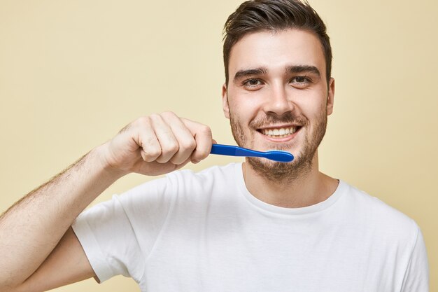 Close-up beeld van positieve jonge ongeschoren man die plastic tandenborstel vasthoudt tijdens het poetsen van tanden in de badkamer voor de spiegel, het verzorgen van mondhygiëne, met tevreden gelaatsuitdrukking