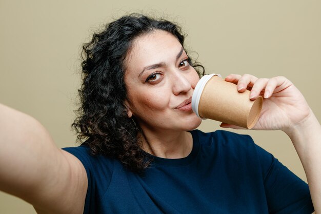 close-up beeld van tevreden vrouw van middelbare leeftijd die een t-shirt draagt met een papieren koffiekopje in de buurt van de mond, kijkend naar de camera die selfie neemt geïsoleerd op een olijfgroene achtergrond