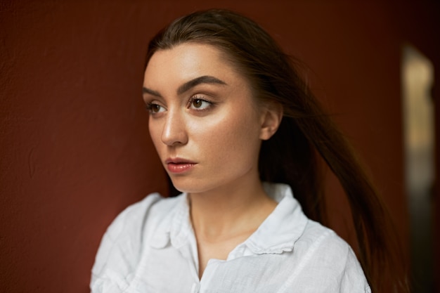 Gratis foto close-up beeld van zelfverzekerde stijlvolle jonge vrouw in wit overhemd lopen tegen bruine muur achtergrond, wind waait haar lange losse haren rond.