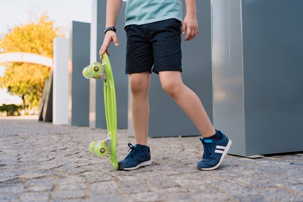 Close-up benen in blauwe sneakers met groene skateboard. Actieve stedelijke levensstijl van jeugd, opleiding, hobby, activiteit. Actieve buitensport voor kinderen. Kind skateboarden.