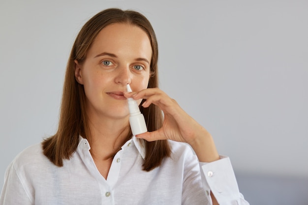 Close-up binnenschot van charmante vrouw die neusspray gebruikt voor loopneus, verkouden wordt, naar de camera kijkt, een wit overhemd draagt, poserend tegen een lichte muur.