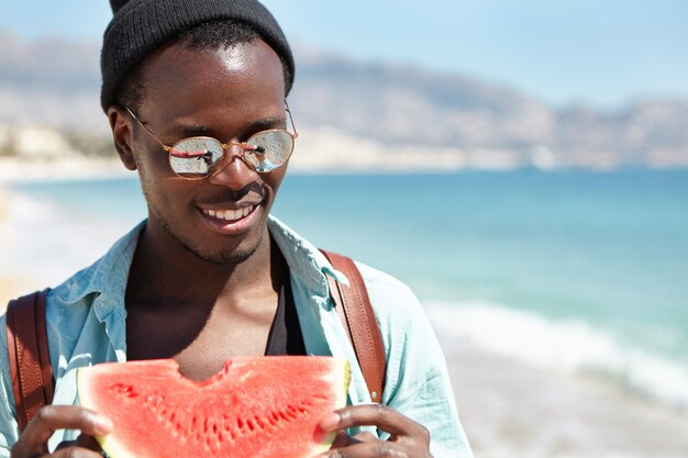 Close-up buiten portret van knappe zorgeloos lachende Afro-Amerikaanse mannelijke reiziger in trendy tinten en hoed wandelen door de zee en het eten van verse rijpe watermeloen