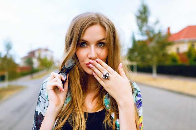 Close-up buiten portret van sensuele hipster blonde vrouw met lange haren en natuurlijke make-up, winderig regenachtig weer, droevige stemming, handen op haar lippen, luchtkus verzenden.