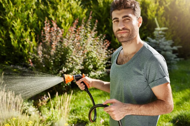 Close-up buitenshuis portret van aantrekkelijke jonge bebaarde Spaanse man in blauw t-shirt met ontspannen gezichtsuitdrukking, planten water geven, bladeren snijden.