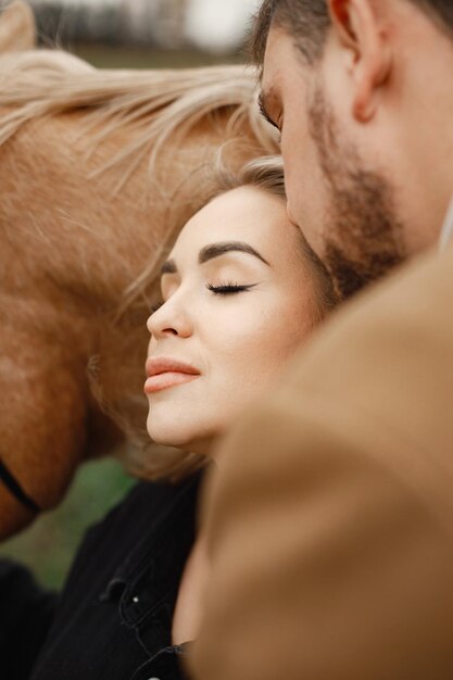 Close-up foto van blonde vrouw, brunette man in het veld met bruin paard. Vrouw met zwarte kleding en man met bruine jas. Man en vrouw knuffelen.