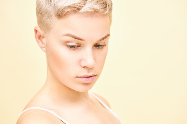 Close-up foto van ernstige doordachte jonge blonde vrouw met stijlvol kort kapsel met peinzende gezichtsuitdrukking. Leuk mooi meisje met perfecte pure huid poseren geïsoleerd, naar beneden te kijken