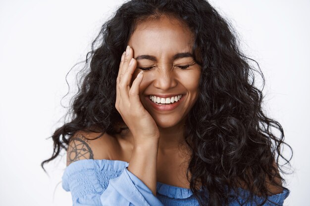 Close-up gelukkige, vrolijke en zorgeloze enthousiaste vrouw met tatoeage in stijlvolle, modieuze blauwe blouse, dichte ogen raken een heldere huid, lachen met plezier, staande witte muur