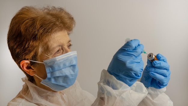 Close-up gezicht van een volwassen vrouwelijke arts met een masker en handen in medische handschoenen, vult een spuit met medicijnen uit een ampul, op een witte achtergrond. een verpleegster houdt een ampul vast. coronavirus.