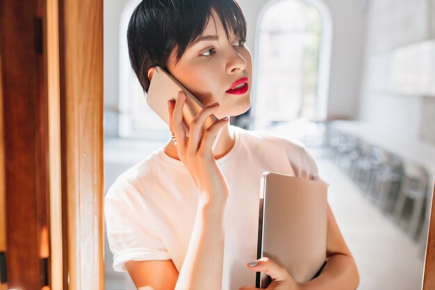 Close-up indoor portret van drukke jonge vrouw met rode lippen en trendy kort kapsel praten over de telefoon