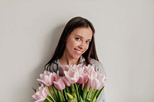 Close-up indoor portret van gelukkig lachend meisje met naakt make-up en lang donker haar poseren met bloemen over witte muur