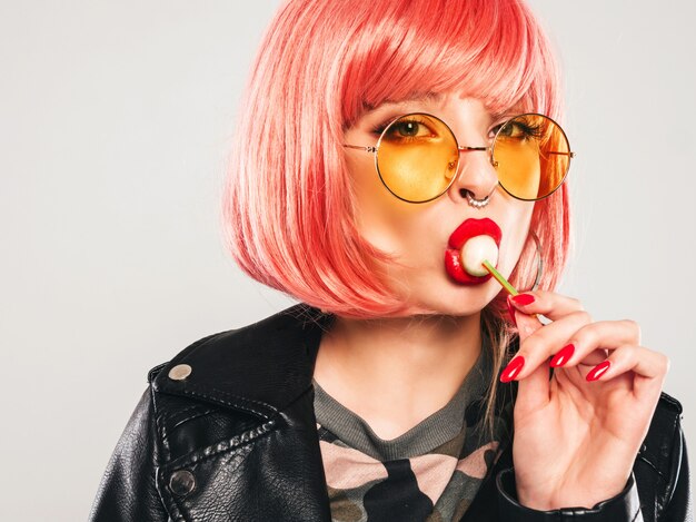 Close-up lippen van jonge mooie hipster slecht meisje in trendy zwarte leren jas en oorbel in haar neus. Sexy zorgeloos lachende vrouw poseren in studio in roze pruik. Positief model likken ronde snoep